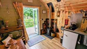 A cozy cabin with a wooden interior, a fireplace, a small kitchen, and a view of the surrounding greenery. - Emlyn's Coppice - Woodland Glamping (Holywell)