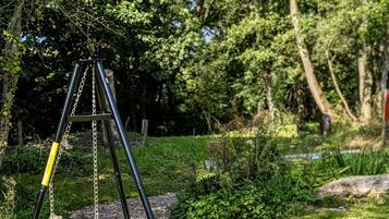 A swing set with a metal frame and a circular seat, positioned on a grassy area with trees and wooden stumps in the background.