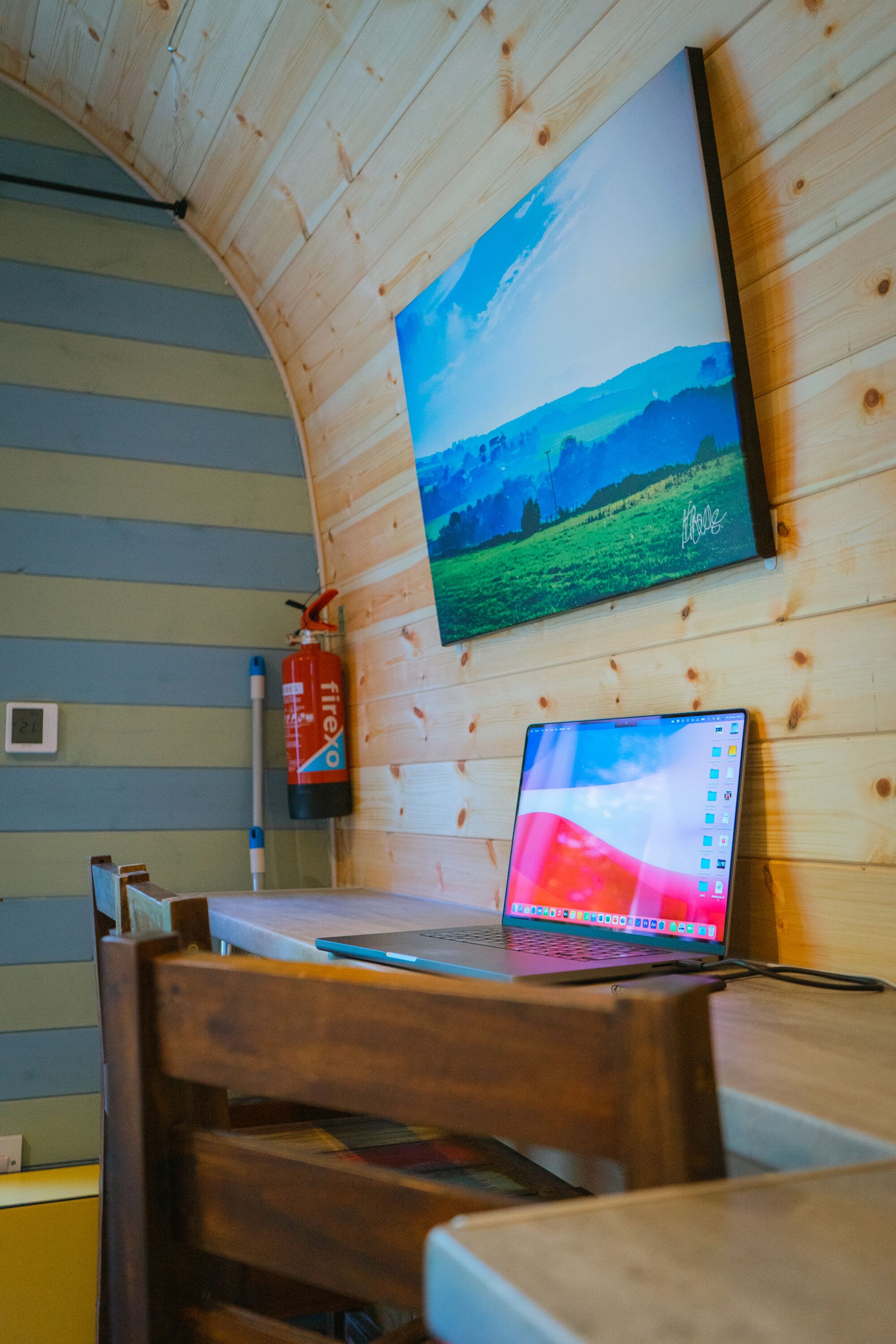 Un interior de madera con una computadora portátil sobre una mesa, un extintor y un cuadro de un paisaje.