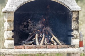 Une cheminée extérieure en pierre, avec un socle en briques, entourée de bûches et de bois en combustion.