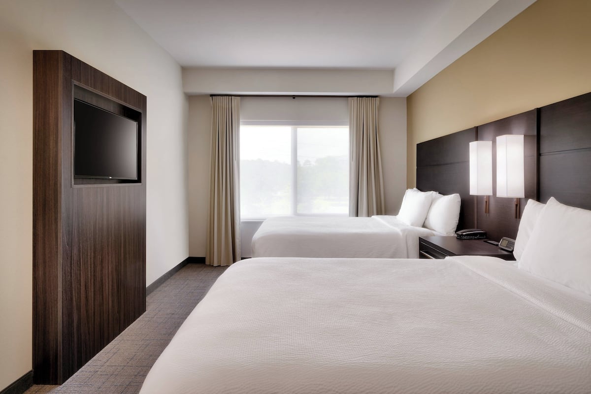 A hotel room with two beds, a flat-screen TV mounted on a wooden panel, a large window with curtains, and a bedside table with a lamp.