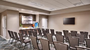 Un salón de conferencias con filas de sillas, un televisor de pantalla plana y una zona de recepción con una planta y un refrigerador para bebidas.