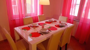 Une table à manger dressée avec des assiettes, des verres et des couverts, entourée de chaises, avec des rideaux roses et une lampe au-dessus.