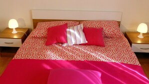 Un lit recouvert d’une couvre-lit à pois rouges et blancs, avec deux oreillers et un coussin rayé. Un tableau représentant des fleurs est accroché au mur au-dessus du lit.