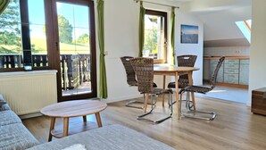 A dining area with a round table and four wicker chairs, a sofa, and a view of a garden through large windows.