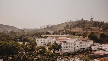 Um edifício grande e branco com vários andares e uma cruz no topo, rodeado por árvores e colinas.