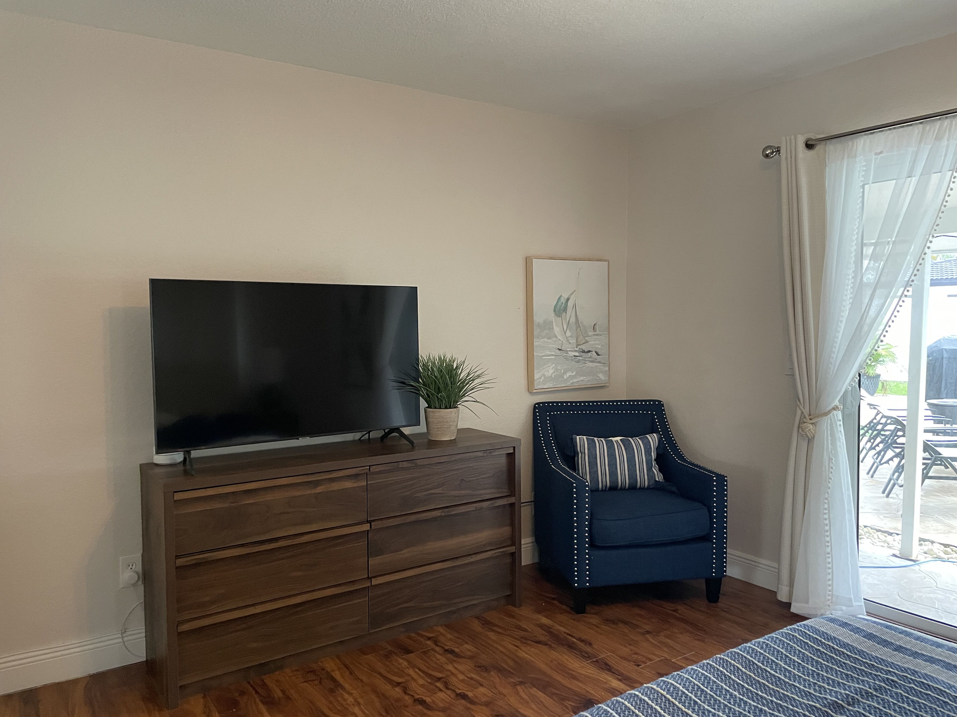 Un salon avec un téléviseur à écran plat posé sur un meuble en bois, une plante en pot, un fauteuil bleu avec un coussin à rayures et un tableau encadré accroché au mur.