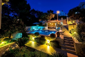 A well-lit outdoor swimming pool area with lounge chairs and a stone wall.
