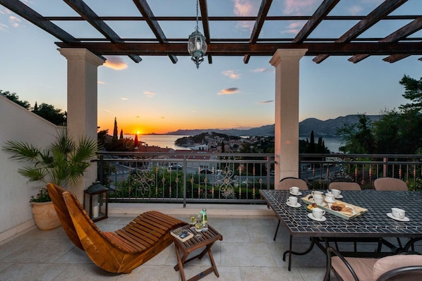 A terrace with a table set for tea, a wooden chair, and a view of the sunset over a city and mountains.