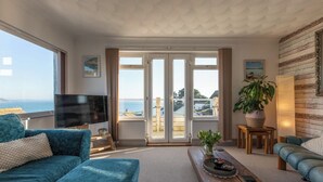 A living room with a large TV, a sofa, a coffee table, and a view of the sea through the sliding doors. - 1 Bed Flat - Sleeps 2 - Pets - Balcony - Parking (Looe)