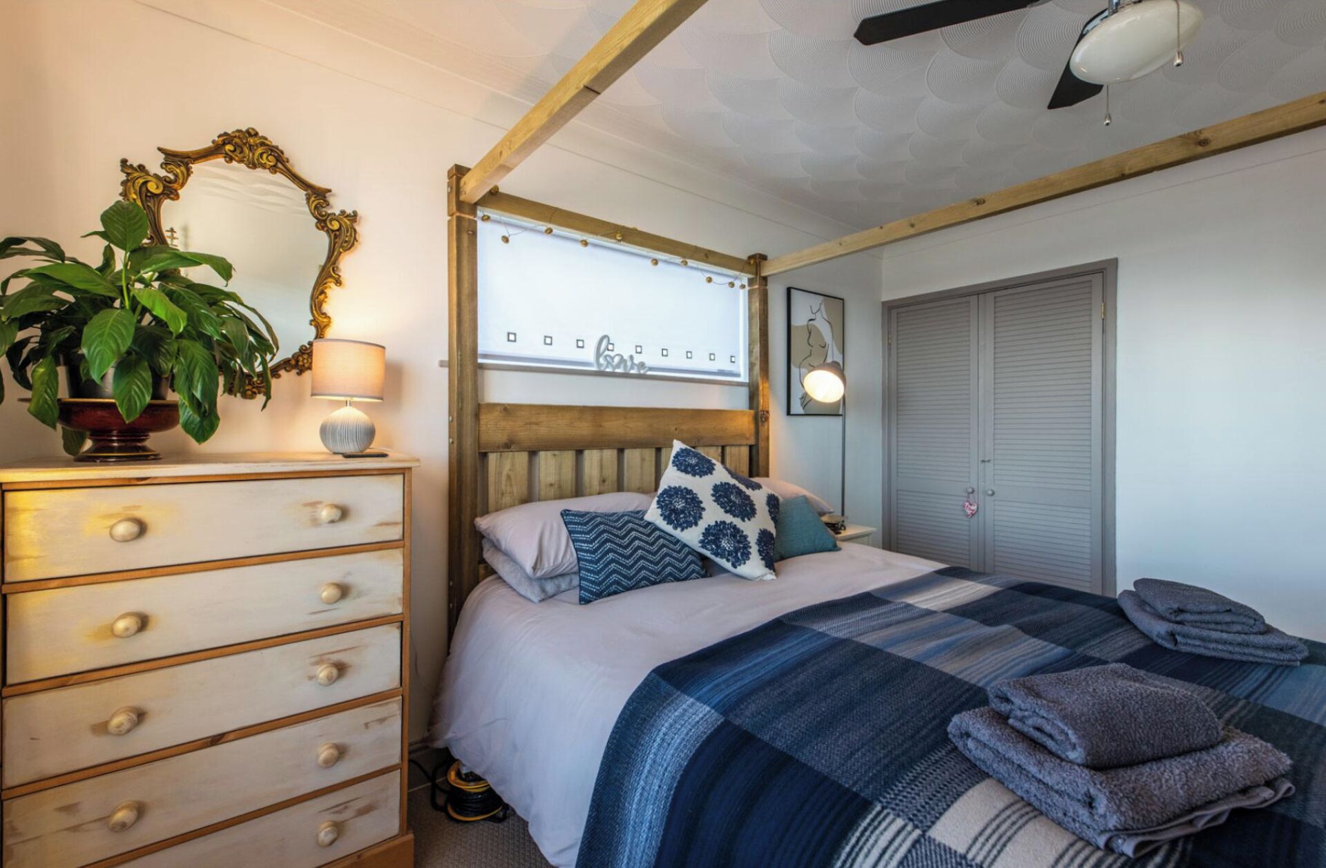 A bedroom with a bed, a dresser, a mirror, a plant, a wall art, and a wardrobe.