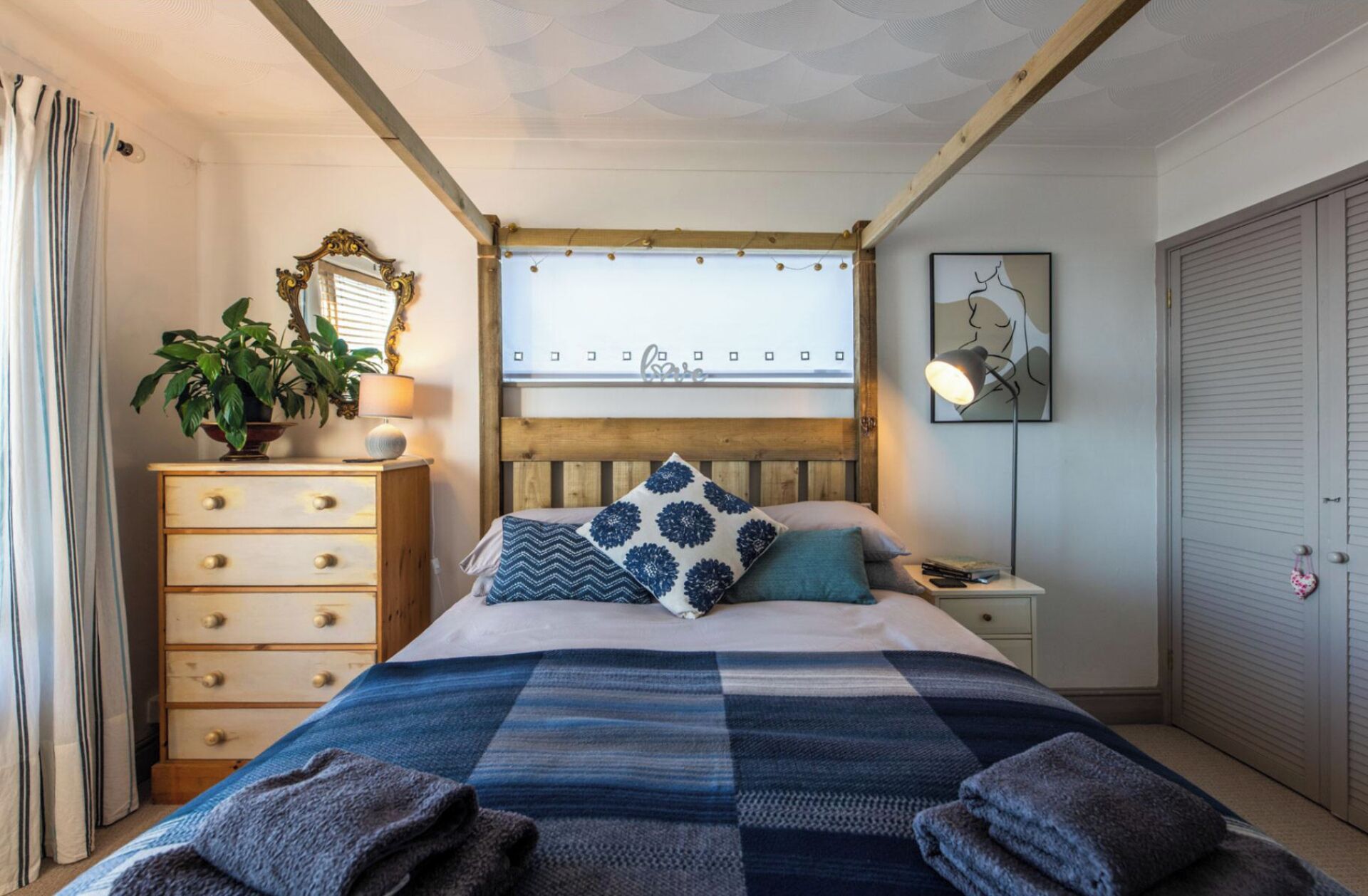 Un dormitorio con cabecera de madera, una cama con manta azul y blanca a cuadros, una mesita de noche con lámpara, un aparador con cajones, una planta en maceta y una ventana con vista al mar.