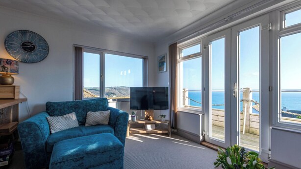 A living room with a blue sofa, a flat-screen TV, and large windows overlooking a coastal view. - 1 Bed Flat - Sleeps 2 - Pets - Balcony - Parking (Looe)