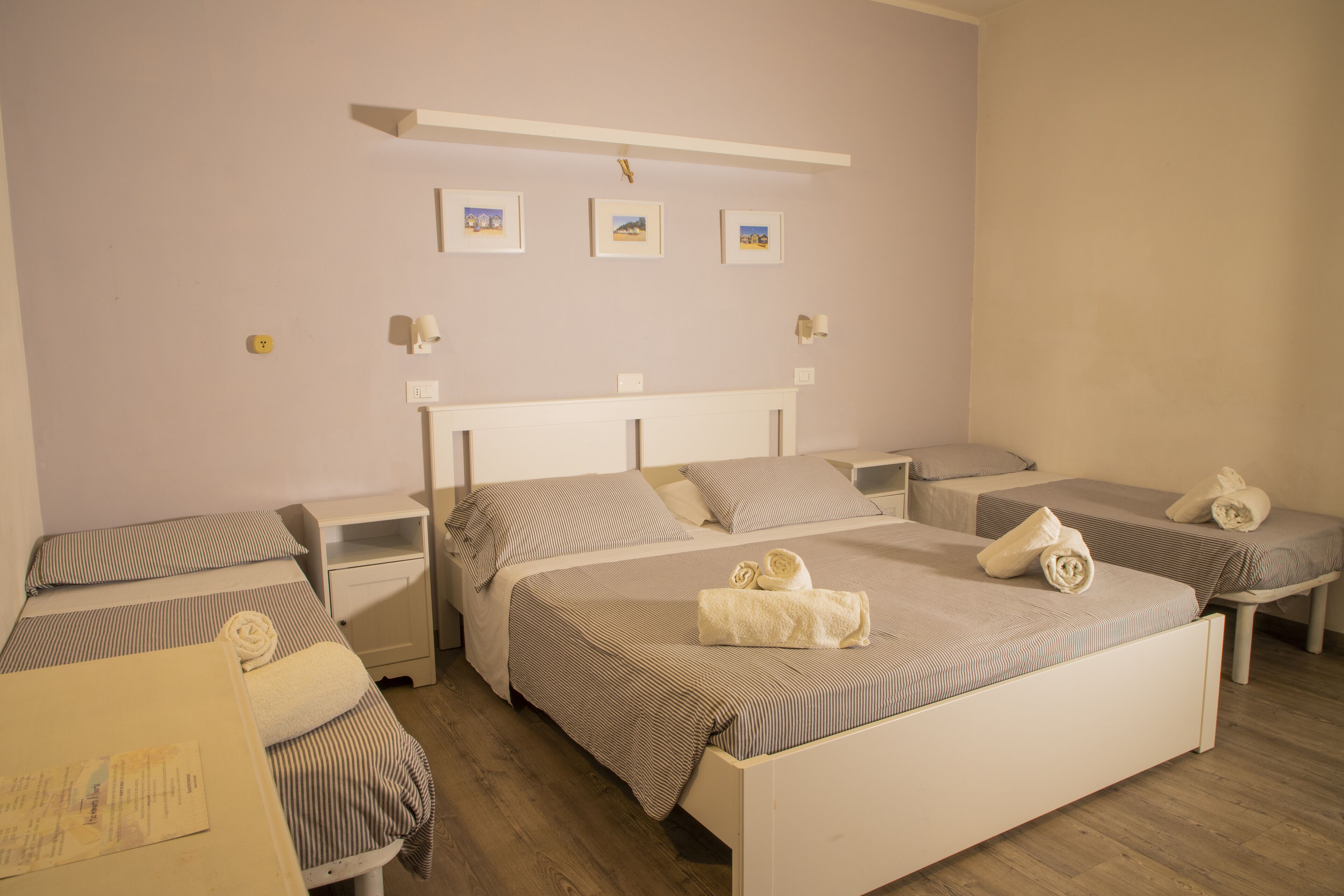 A hotel room with two single beds, a large bed, a nightstand, a bedside table, and a wall-mounted shelf with pictures.