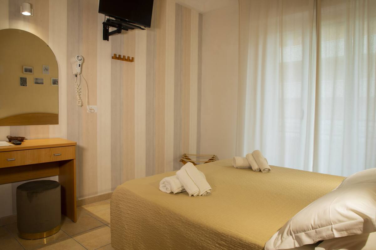 Habitación de hotel con cama, escritorio de madera, espejo, secador de pelo y televisión montada en la pared.