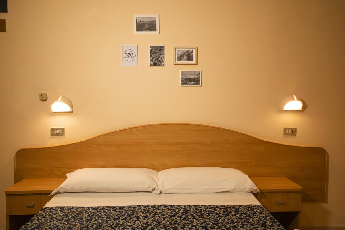 Una cama bien hecha con cabecera de madera, dos mesitas de noche con lámparas, y cuatro cuadros enmarcados en la pared.