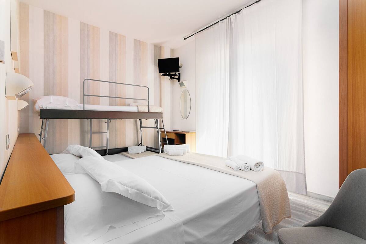 Habitación de hotel moderna con literas, un escritorio con lámpara, una mesita de noche, una silla, un televisor montado en la pared y un gran ventanal con cortinas.