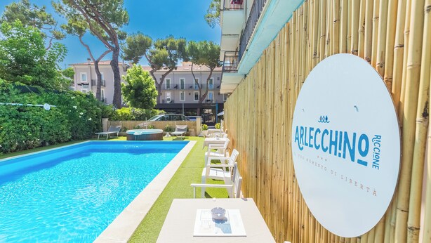 Une piscine entourée d’une clôture en bambou, avec des chaises longues et un panneau indiquant « ARLECCINO RICCIONE ».