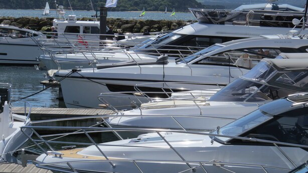 Un port de plaisance où sont amarrés plusieurs yachts, une côte rocheuse en arrière-plan et un drapeau flottant au sommet d’un mât.