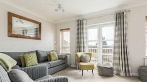 Un salon avec un canapé gris, un fauteuil jaune et un fauteuil à rayures. Il y a un grand miroir au mur et une fenêtre avec des rideaux.