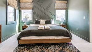 Un dormitorio con una cama grande, dos mesitas de noche, una lámpara colgante y dos ventanas con persianas.