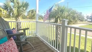 A covered porch with a wooden floor, a white railing, and a view of palm trees and a flag. - 2 Beach view Cottages, Perfect family get together (Gulfport)