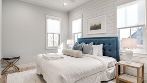 Un dormitorio con una cama grande, ropa de cama blanca, un cabecero azul y una mesita de noche de madera con una lámpara.