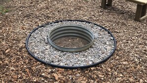 Un foyer circulaire, constitué d’un anneau métallique et rempli de gravier, situé dans une zone boisée.