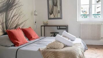 Une chambre avec un grand lit, un luminaire suspendu, un radiateur et une fenêtre donnant sur des immeubles.