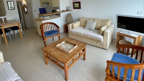 Sala de estar con sofá, dos sillas de madera, una mesa de centro, televisor y zona de cocina al fondo.