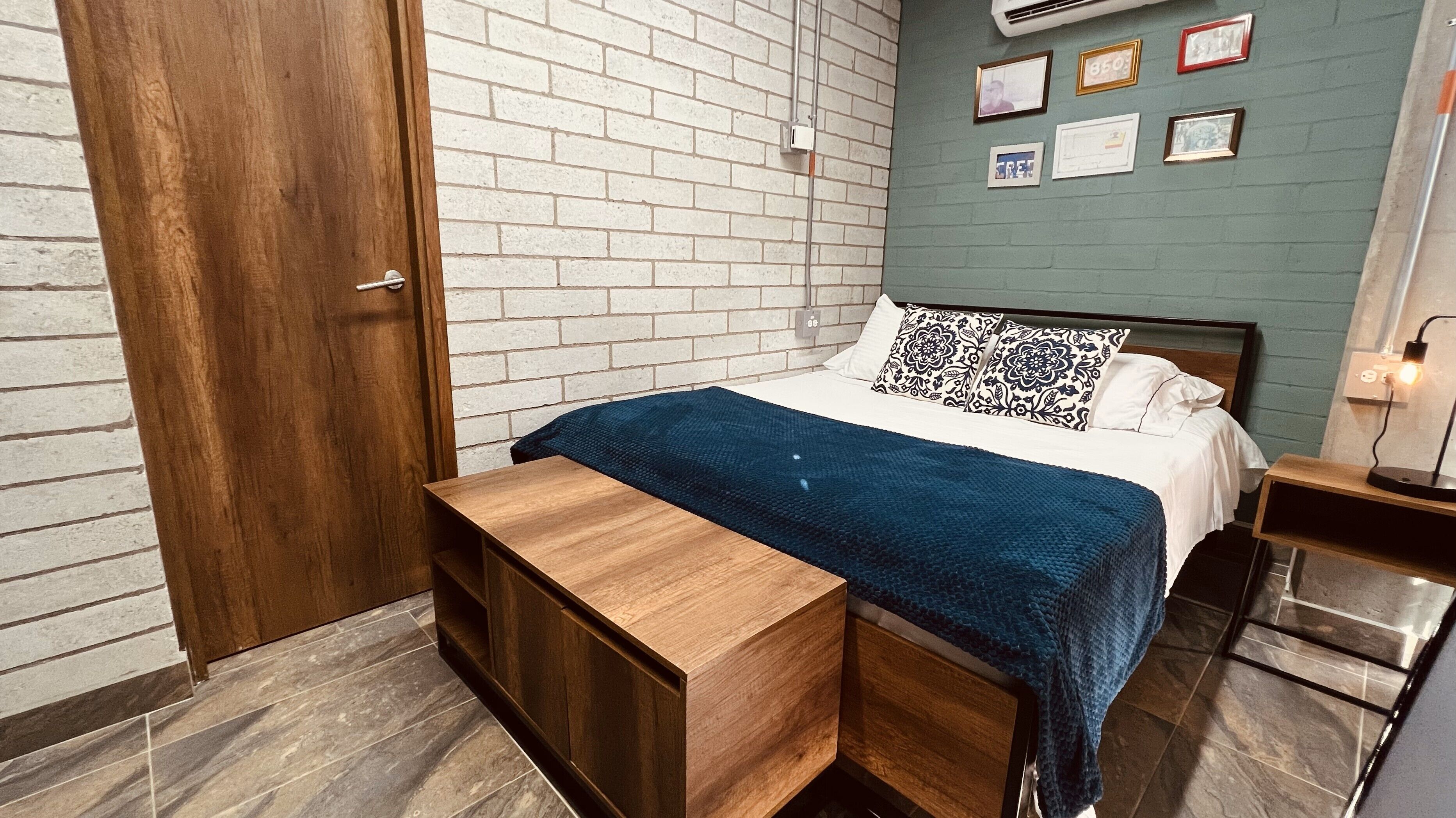 Une chambre avec un lit en bois, un banc, un mur en briques et une petite table de chevet avec une lampe.