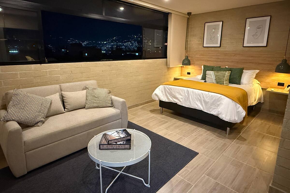 Un dormitorio moderno con vista a la ciudad, un sofá, una cama con una manta amarilla y dos lámparas de noche.