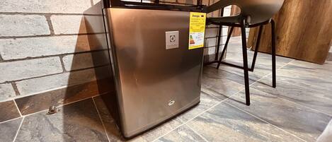 A compact, metallic-colored refrigerator with a sticker indicating energy efficiency, placed on a tiled floor with a brick wall and a chair in the background.