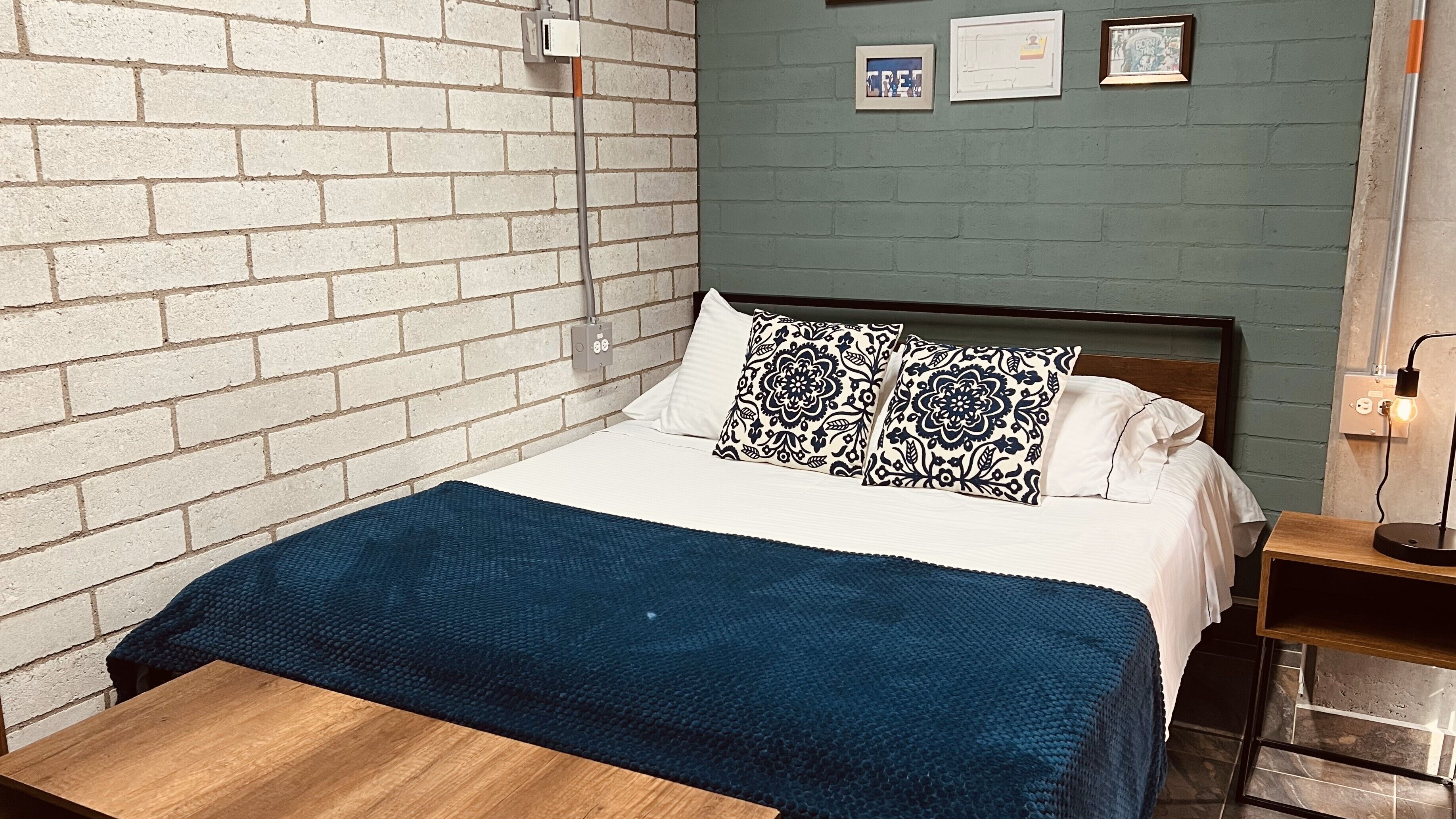 Een kamer met een bed, een houten nachtkastje, een blauwe deken, een bakstenen muur en een groene muur met ingelijste foto's.