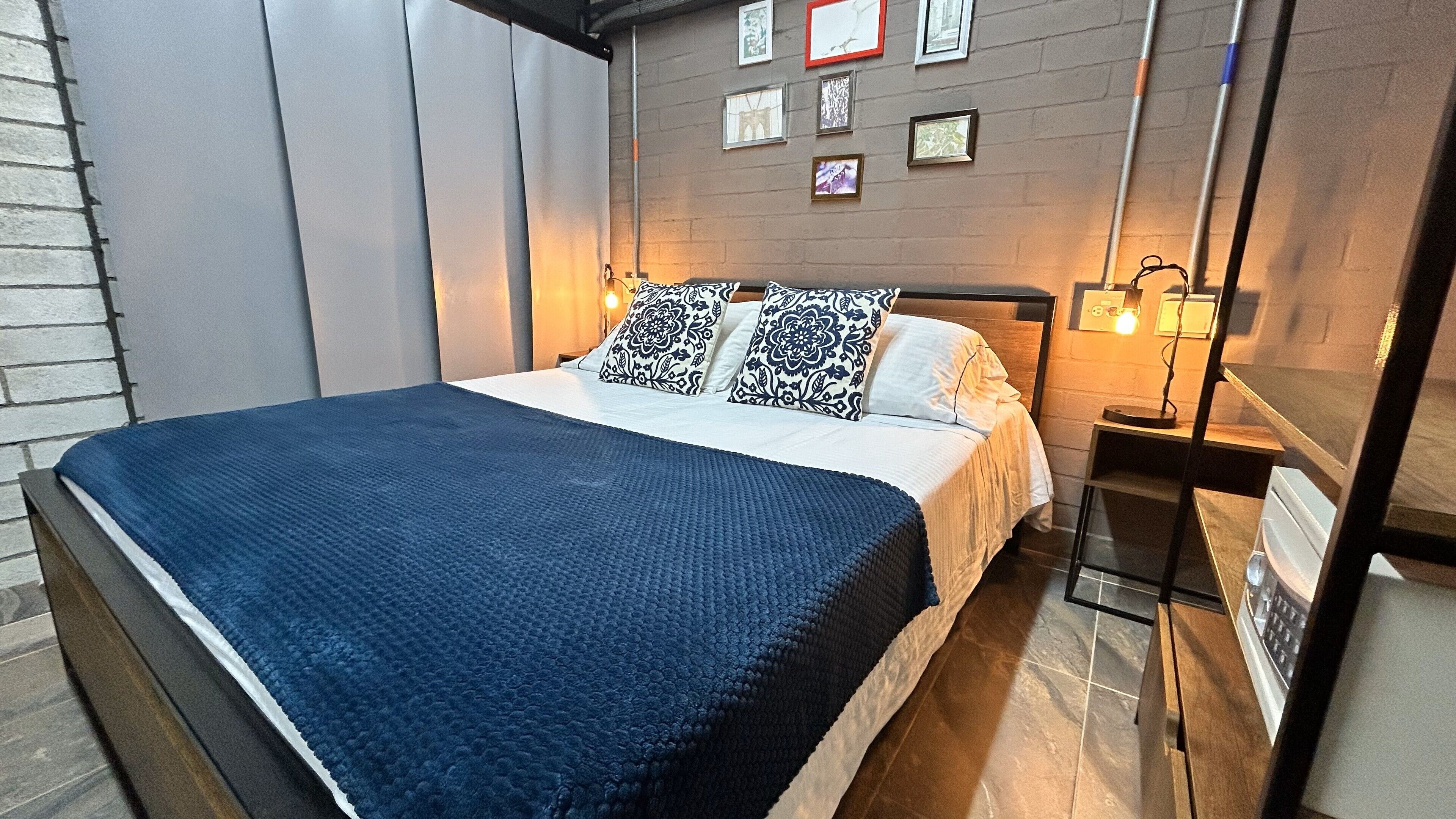 A modern bedroom with a large bed, a bedside table with a lamp, and a wall with framed pictures.