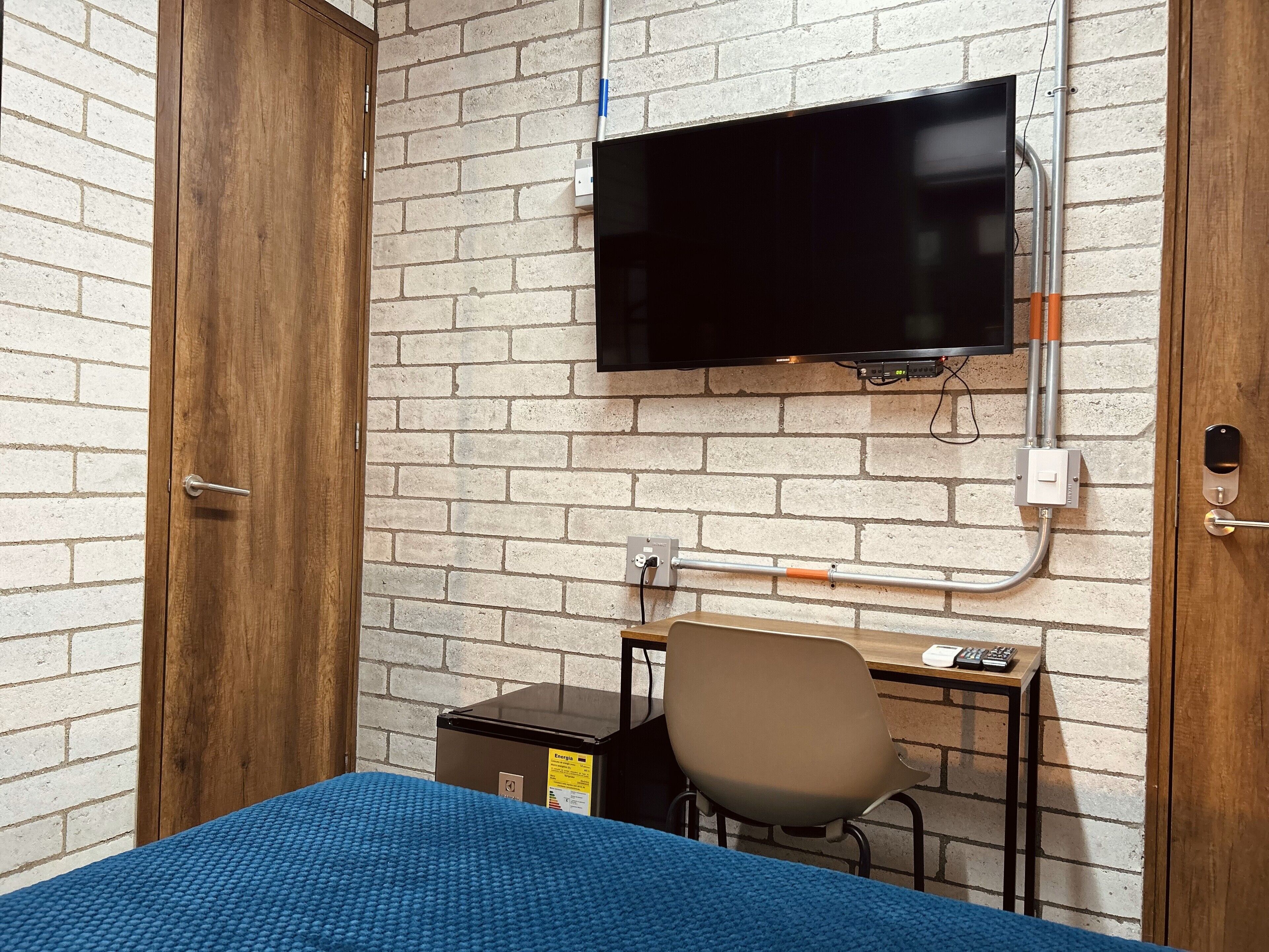 A room with a brick wall, a mounted TV, a desk with a chair, and a bed with a blue blanket.