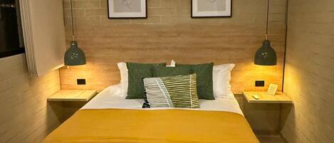 A hotel room with a bed, two bedside tables, a nightstand, a wall-mounted air conditioner, and two framed artworks.