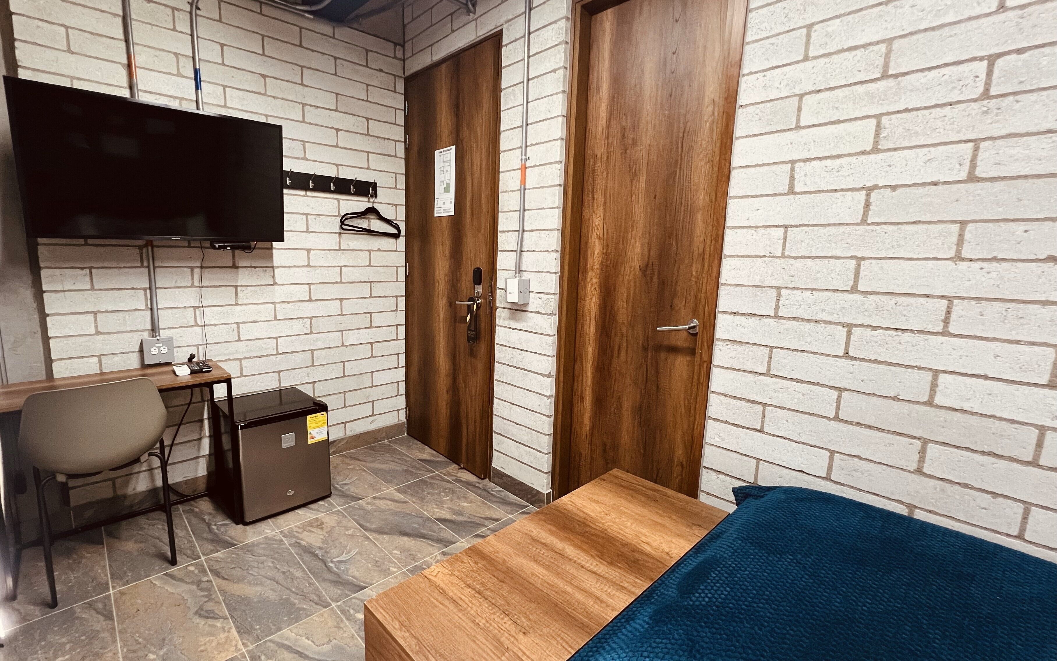 Una stanza piccola con un letto, una scrivania, una sedia, una TV fissata al muro e un armadio in legno.