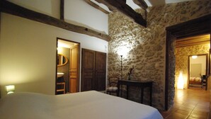 Un dormitorio con una cama grande, vigas de madera, paredes de piedra, una mesita con una lámpara y un baño visible a través de una puerta abierta.