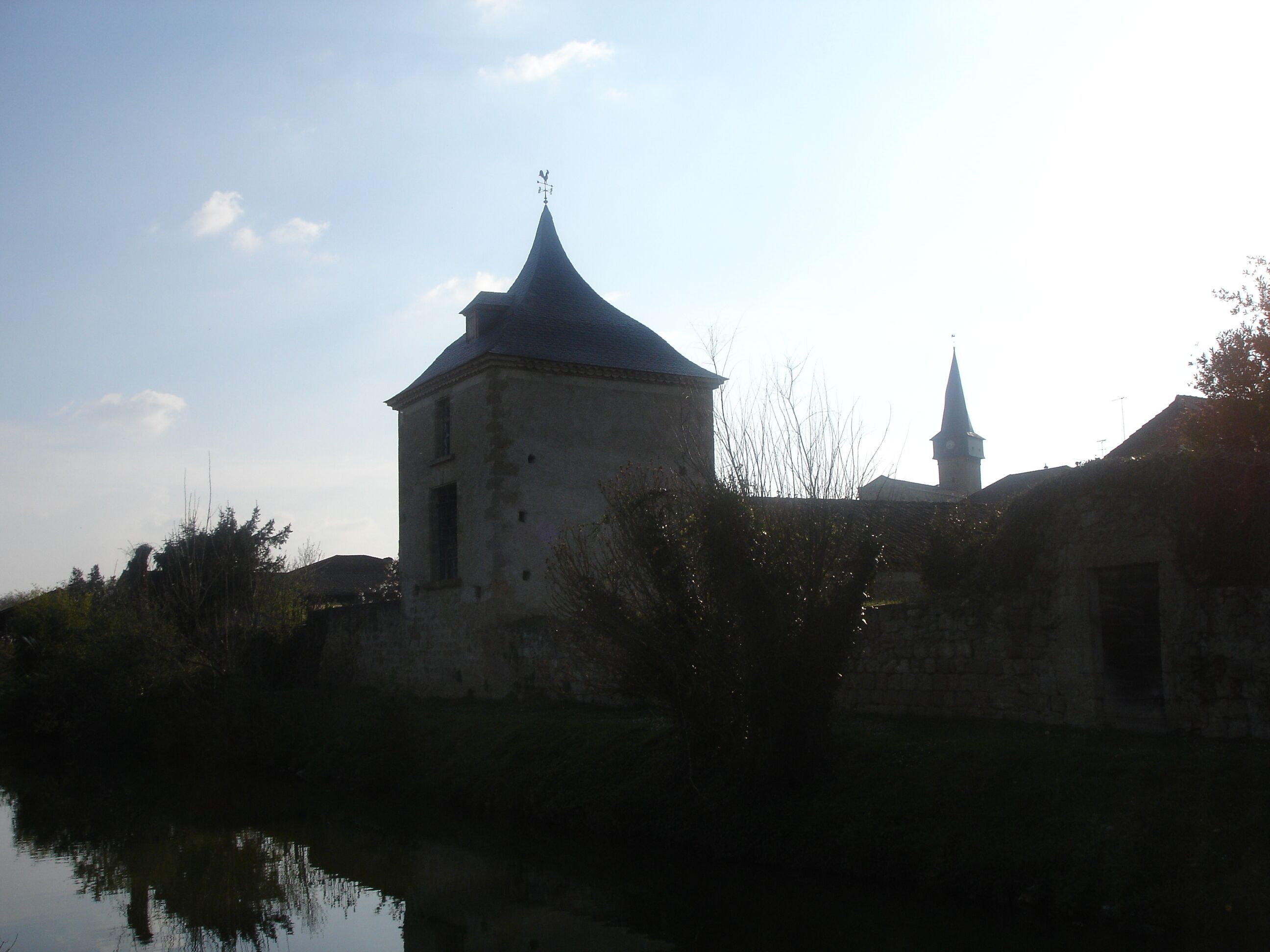 Ein historisches Gebäude mit einem markanten Turm und einem kleineren, daran angebauten Bauwerk, umgeben von Vegetation und einem Gewässer.