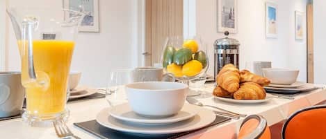 Una mesa de desayuno con una jarra de jugo de naranja, un bol de fruta, croissants y una cafetera.