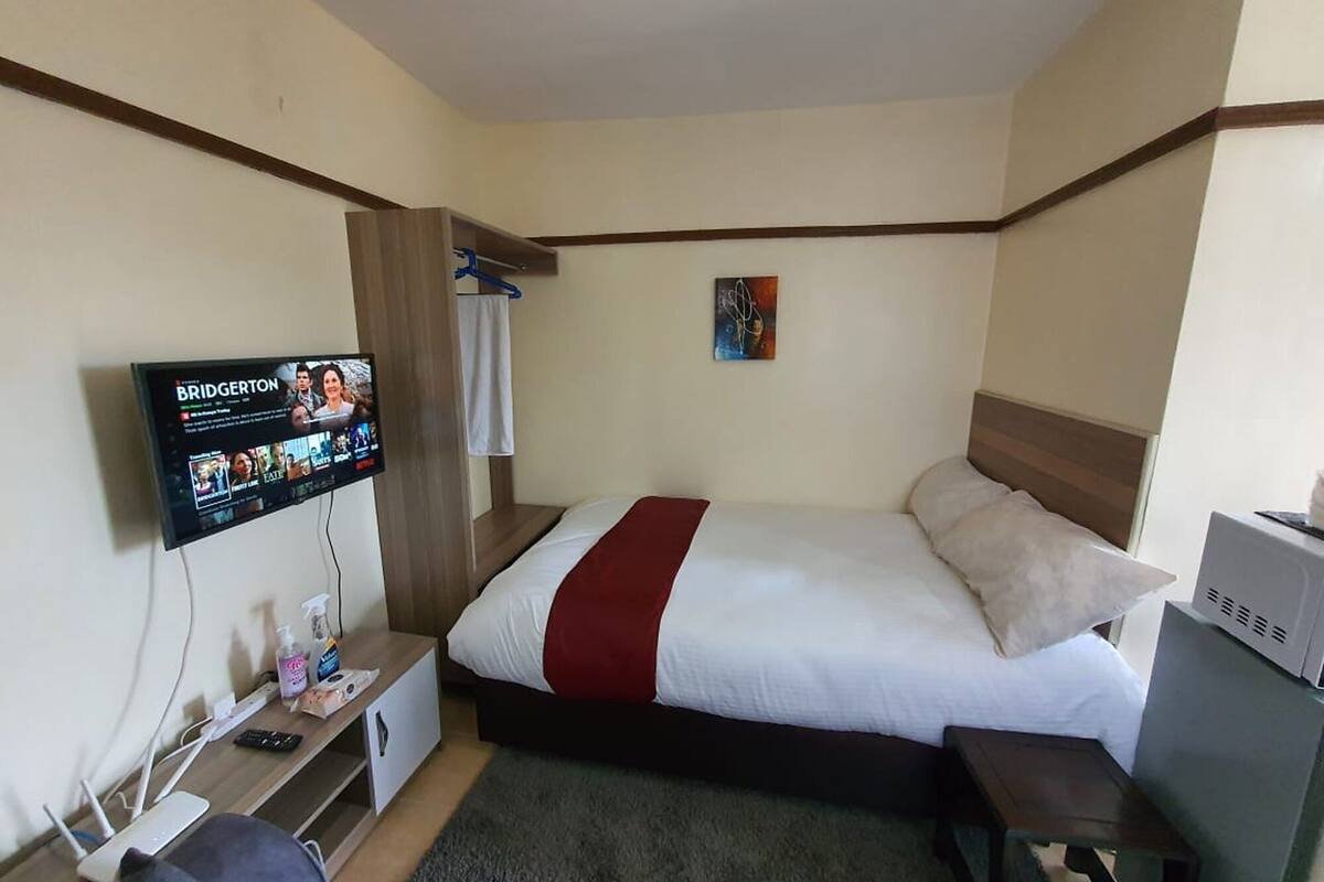 A hotel room with a bed, a TV mounted on the wall, a desk with a computer, and a small table.