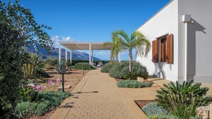 A pathway leading to a white building with wooden shutters, surrounded by landscaped gardens and palm trees. - Villa Etesiae (Castellammare del Golfo)