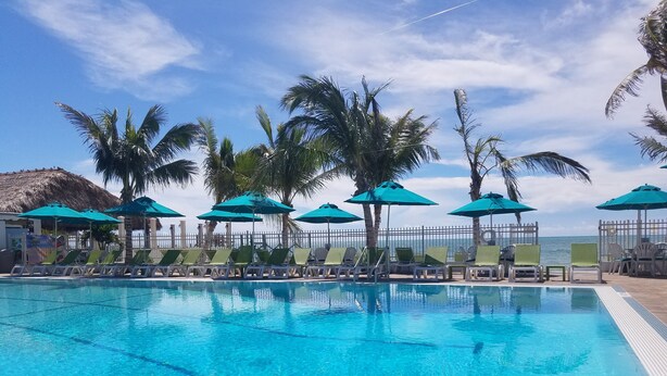 A swimming pool with lounge chairs and umbrellas, surrounded by palm trees and a view of the ocean. - Beautiful waterfront 3/2 home with dock and boat lift. Cabana Club included! (Key Colony Beach)