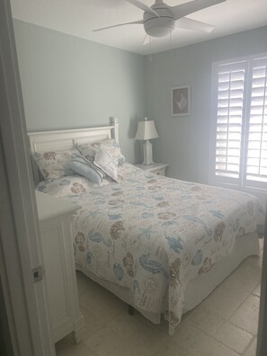 A bedroom with a bed, a nightstand, a ceiling fan, a window with blinds, and a wall-mounted artwork.