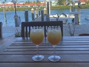 Two glasses of a yellow beverage on a wooden table with a view of a waterfront and buildings in the background.