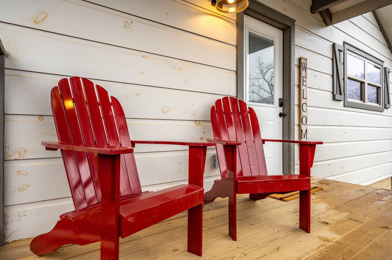 Två röda Adirondack-stolar står på ett trädäck utanför en vit byggnad, och en skylt med texten ”VÄLKOMMEN” hänger på byggnaden.