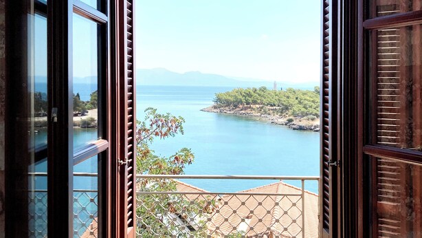 An open door leading to a balcony with a view of a coastal landscape, including a body of water and distant mountains.
