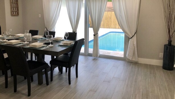 A dining area with a table set for four, large windows with white curtains, and a view of a swimming pool. - 3-Bedroom Heated Private Pool Vacation Home just minutes from the Beach Pet (Deerfield Beach)