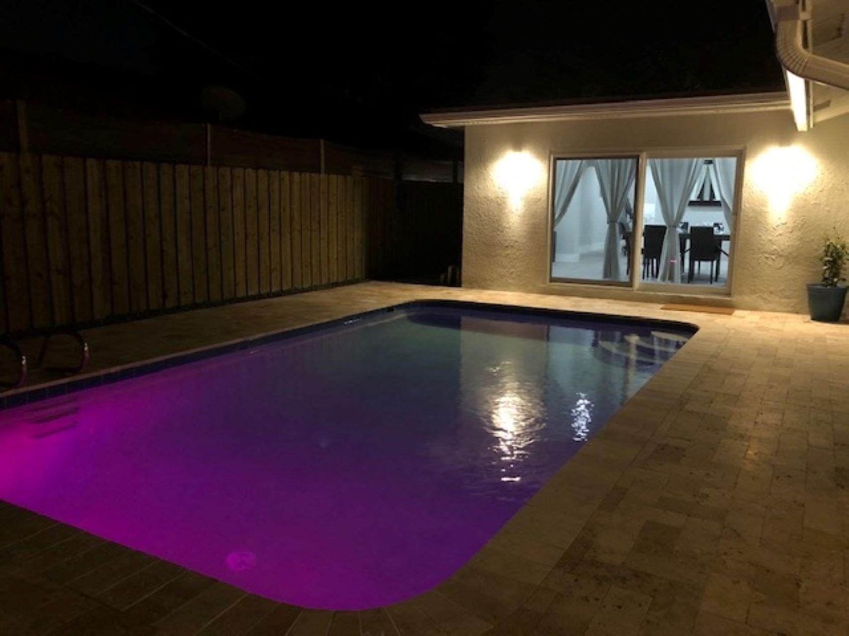 Ein Pool mit violetter Beleuchtung in der Nacht, umgeben von einem Holzzaun und einem Haus mit Glastür und Vorhängen.